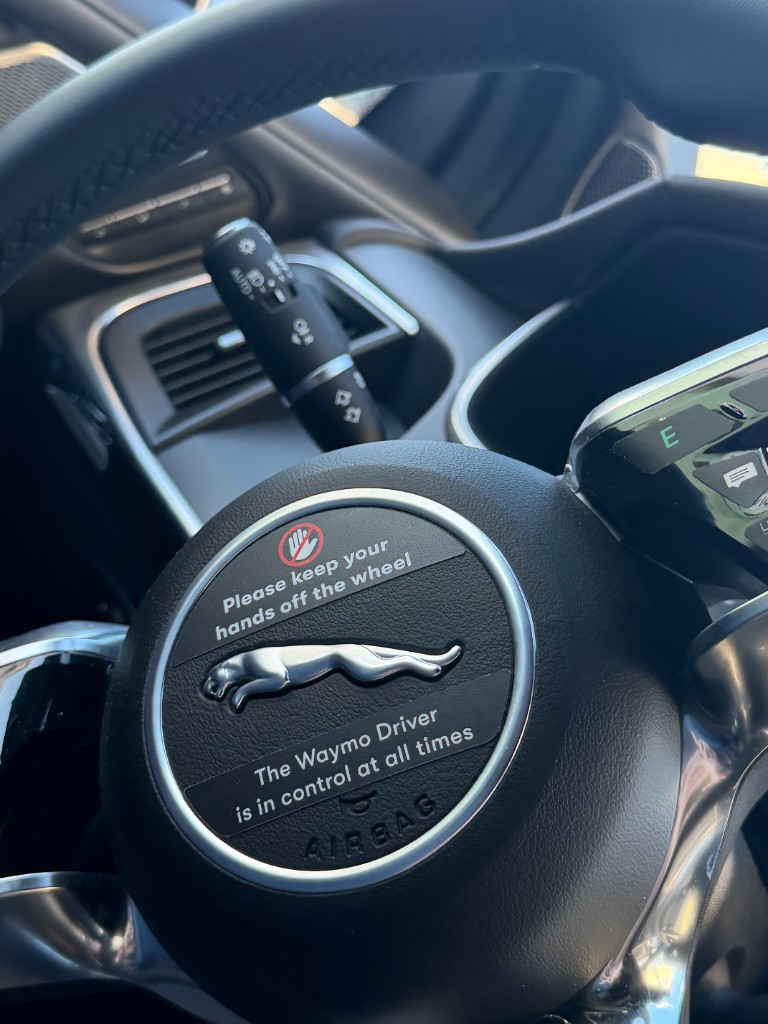 Inside a Waymo Jaguar with a steering wheel label reading that the Waymo Driver is in control at all times.